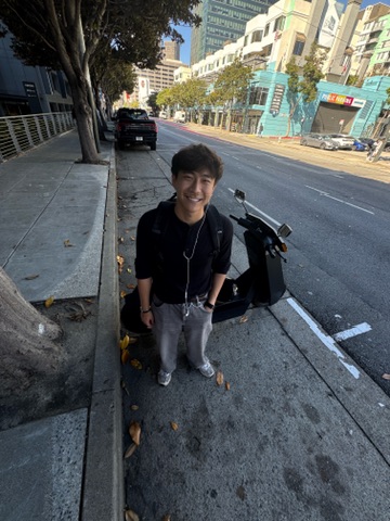 Daniel standing on a San Francisco sidewalk beside a scooter